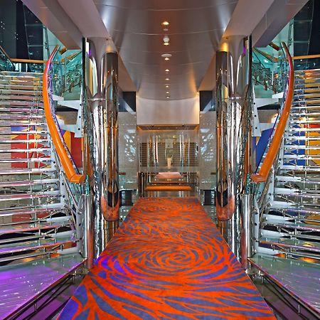 Golden Sands Boutique Hotel-Dubai Creek ภายใน รูปภาพ The photo shows a modern, elegantly designed interior space, likely on a cruise ship. It features two sweeping, curved staircases made of glass and metal, flanking either side of a central corridor. The ground is covered with a vibrant orange carpet 