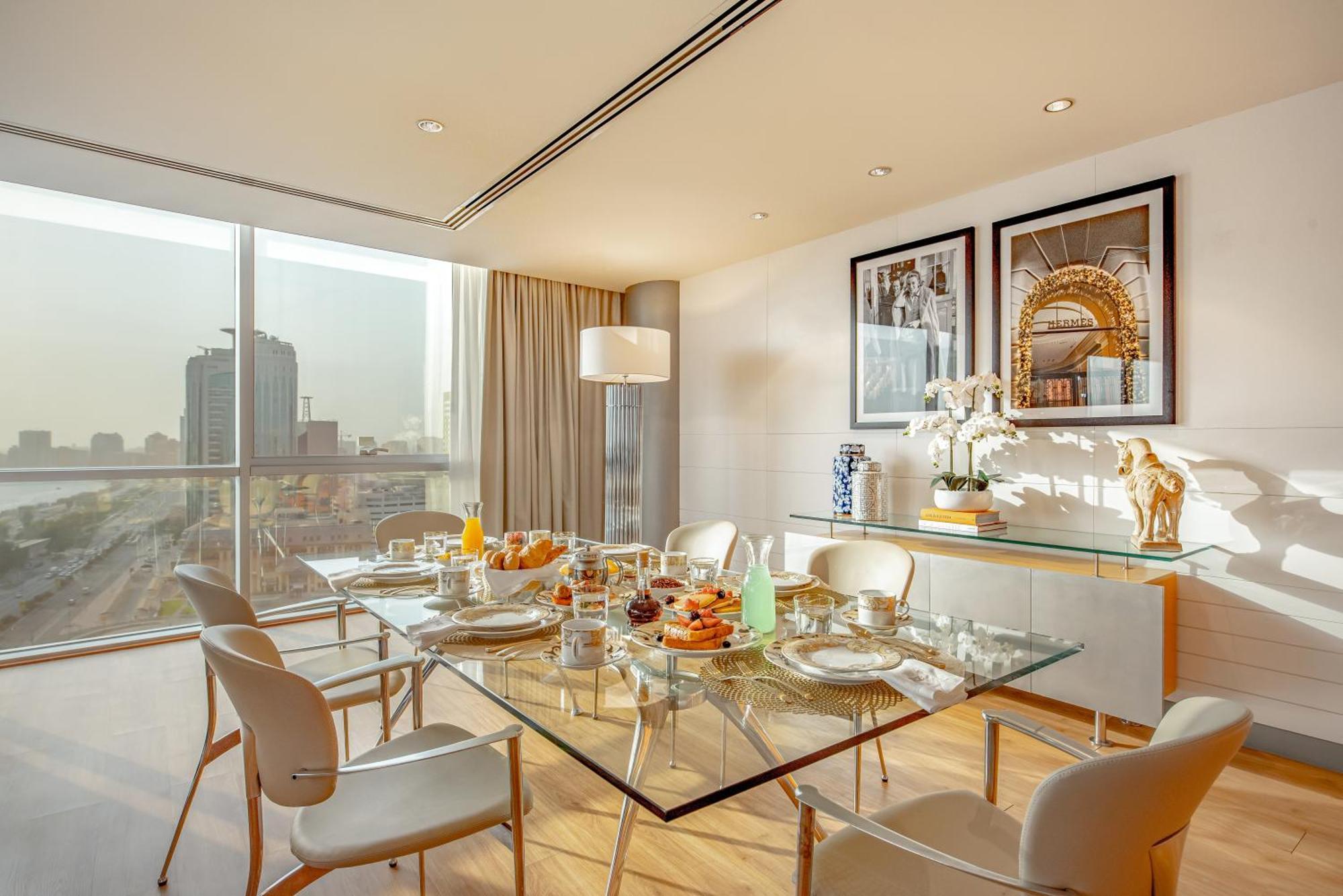 Golden Sands Boutique Hotel-Dubai Creek ห้อง รูปภาพ The photo shows a modern dining room featuring a glass dining table set for a meal. The table is adorned with plates, cutlery, glasses, and a variety of food items, suggesting a breakfast or brunch setting. Surrounding the table are sleek, white chai