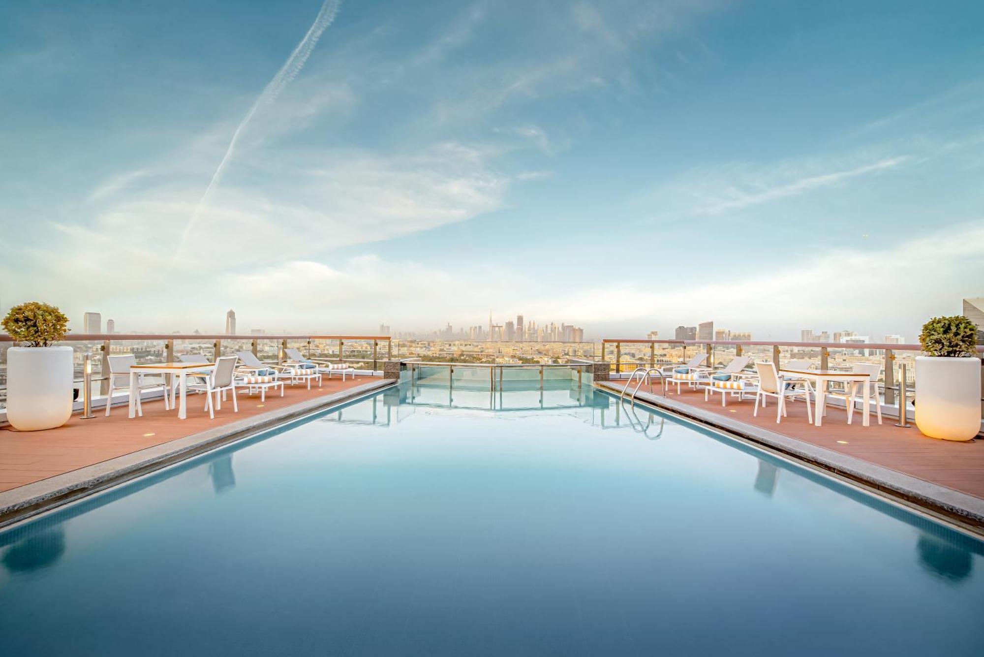 Golden Sands Boutique Hotel-Dubai Creek ภายนอก รูปภาพ The photo showcases a rooftop swimming pool with a stunning view of a city skyline in the background. The pool is calm and reflects the sky, which is clear with a few clouds. Surrounding the pool are several white lounge chairs arranged for relaxatio