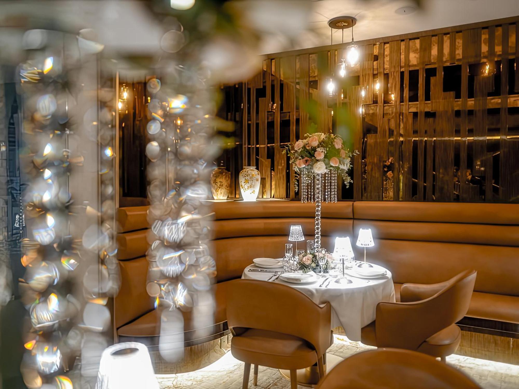 Golden Sands Boutique Hotel-Dubai Creek ภายนอก รูปภาพ The photo depicts an elegant restaurant interior. There is a round dining table set for two, complete with a white tablecloth, center floral arrangement, and small lamps. The seating consists of curved, brown leather chairs, suggesting a cozy atmosph