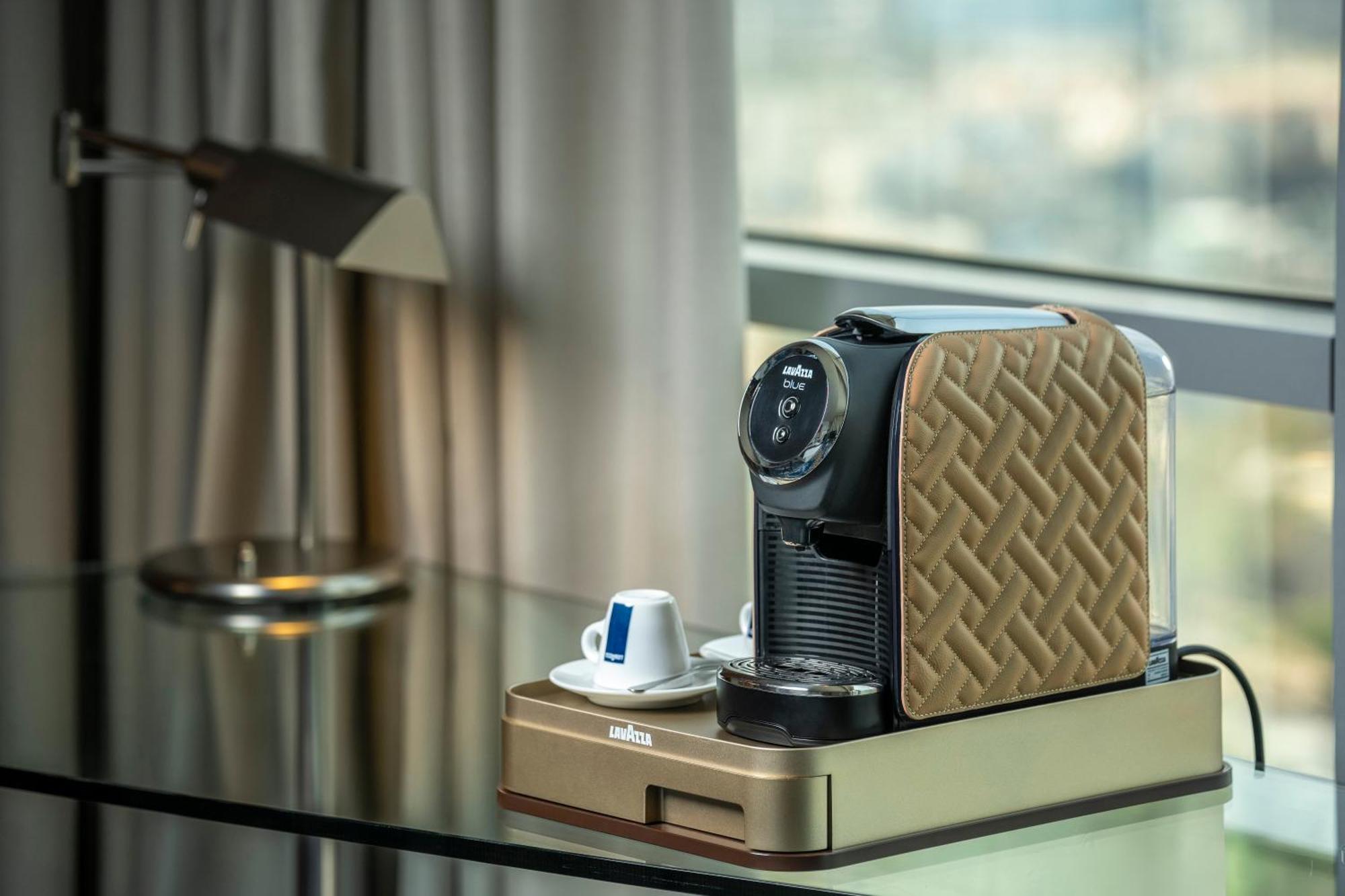Golden Sands Boutique Hotel-Dubai Creek ห้อง รูปภาพ The photo features a coffee machine placed on a sleek table. The machine has a stylish design with a patterned brown exterior. Next to the machine, there is a small saucer with a blue and white coffee cup. The background includes a large window with 